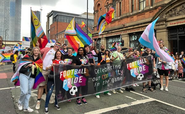 A group of people stand behind a football pride banner waving rainbow flags